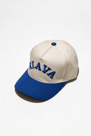 A baseball cap with an off-white crown and a royal blue brim, featuring the word 'KILAVA' embroidered in blue arched letters across the front. The cap is photographed on a clean, light off-white background with a soft shadow.