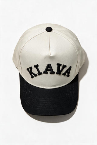 A baseball cap with an off-white crown and a black brim, featuring the word 'KIAVA' embroidered in black arched letters across the front. The cap is photographed from a top-down angle on a clean white background.