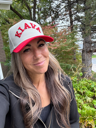 A woman wearing a white and red baseball cap with 'KIAVA' embroidered in red across the front, photographed outdoors in front of trees and shrubs.