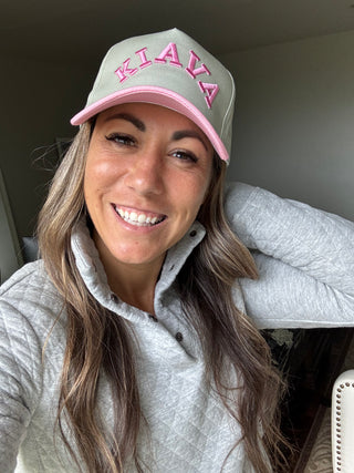 A woman wearing a light gray baseball cap with pink trim and pink embroidered lettering that reads 'KIAVA', paired with a light gray pullover sweatshirt in an indoor setting.