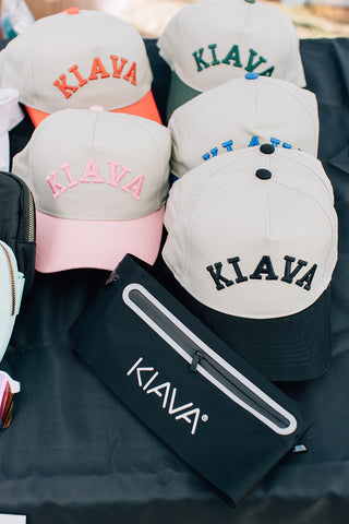 Several baseball caps and a black pouch with the brand name KIVA are arranged on a black cloth. The caps are light gray with embroidered KIVA logos in orange, green, pink, blue, and black, each with a different colored brim.